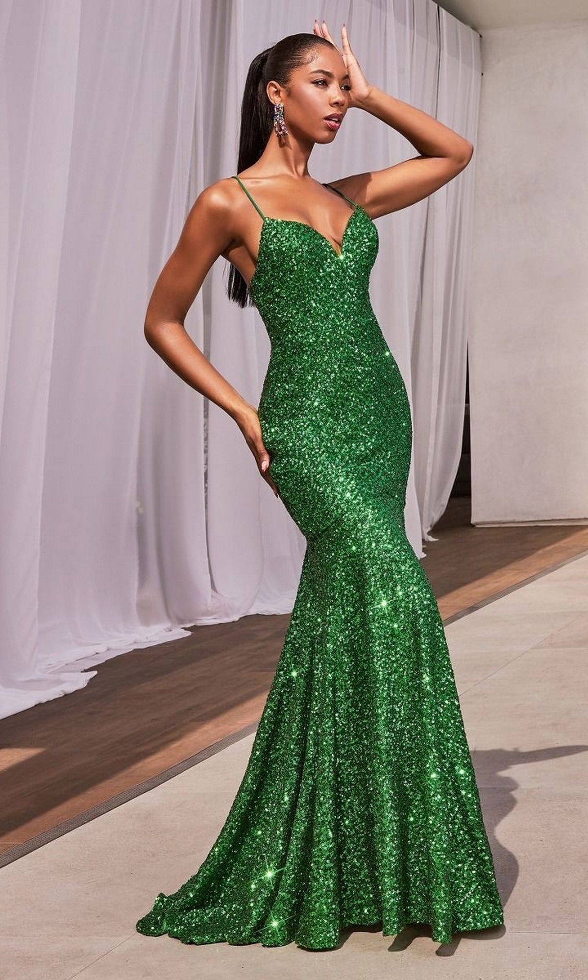 A woman poses outdoors near white drapes and a modern building, wearing the Lace-Up Open-Back Long Sequin Prom Dress CH066—a fitted, floor-length green gown with thin straps.